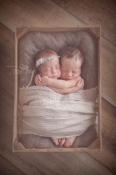 two babies are sleeping in a box on a wooden floor with their arms around each other