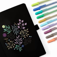 several markers and pens are next to a notebook with an image of flowers on it