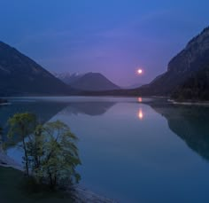 the moon is setting over a lake with mountains in the background