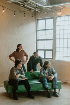 four people sitting on a green couch in an empty room with exposed ceiling and windows