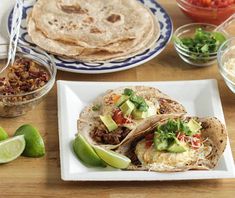 three tacos on a white plate with limes and other ingredients around them,