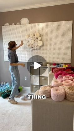 a woman standing in front of a wall with yarn on it and balls of yarn behind her