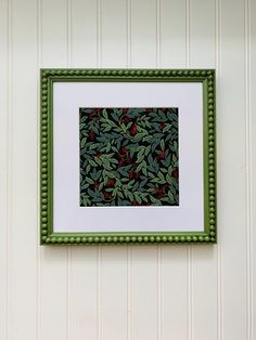 a green frame hanging on the wall with red berries and leaves in front of it