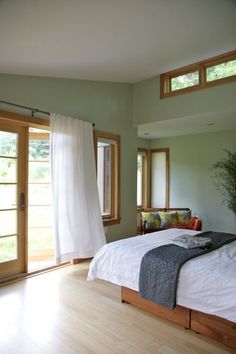 a bed sitting in a bedroom next to two windows and a plant on top of it