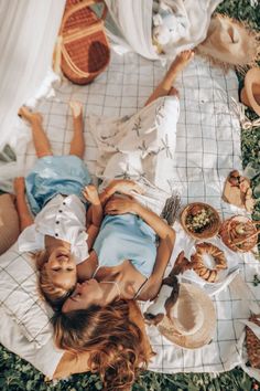 two women laying on a blanket in the grass with food and hats around them,