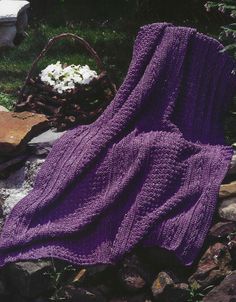 a purple knitted blanket sitting on top of a pile of rocks next to flowers