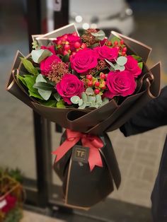 a bouquet of pink roses wrapped in brown paper with red ribbon and green leaves is held by someone's hand