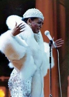 a woman in a white fur coat standing next to a microphone and holding her hands out