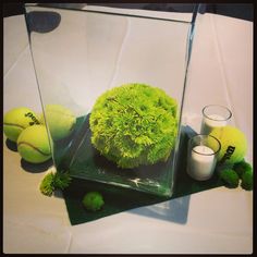 a glass vase filled with green plants and tennis balls on top of a white table cloth