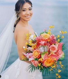 a woman in a wedding dress holding a bridal bouquet by the water with an ocean behind her