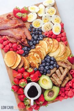 an assortment of breakfast foods laid out on a cutting board with fruit, eggs, bacon, pancakes and avocado