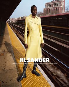 a woman in a yellow coat stands on a train platform with her hands in her pockets