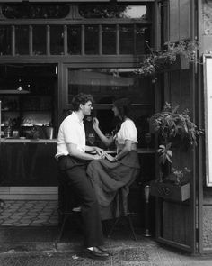 two people sitting at a table in front of a store with plants on the windows