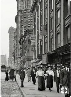 black and white photograph of people walking on the sidewalk in an old city street with tall buildings