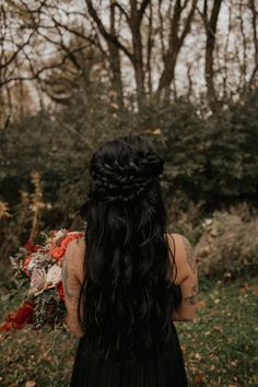 a woman with long black hair standing in front of some trees and flowers, looking at the sky