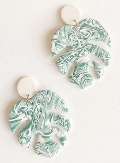 two green and white leaf shaped earrings on a white surface, one is hanging from the ear