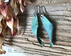 two blue and green fish shaped earrings hanging from hooks on a wooden wall next to flowers