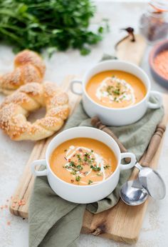 two bowls of carrot soup on a cutting board with pretzels in the background