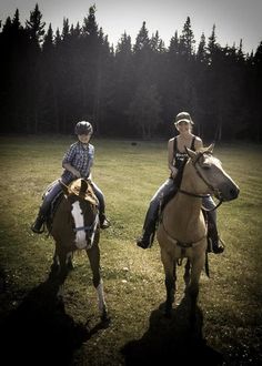 two people riding on the backs of horses in a grassy field with trees behind them