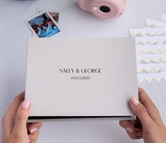 two hands holding a wedding album on top of a table with photos and other items