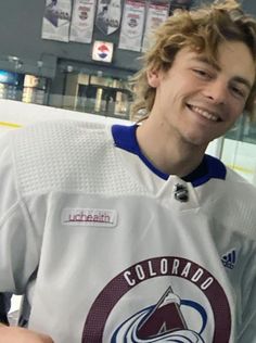 a man with long hair wearing a hockey jersey and smiling at the camera while standing in front of an ice rink