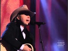 a man wearing a cowboy hat and holding a guitar in front of a microphone on stage