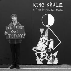 a man holding a sign standing next to a wall with graffiti on it that reads king krule 6 feet beneath the moon