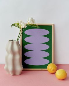 two lemons sit next to a white vase with flowers in it on a pink surface