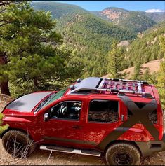 a red pick up truck parked on top of a dirt road next to trees and mountains