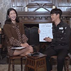 a man and woman sitting next to each other in front of a fireplace holding up a piece of paper