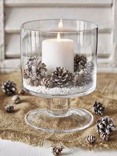 a lit candle in a glass bowl filled with pine cones and snowflakes