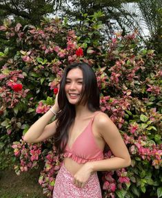 a woman standing next to a bush with pink flowers in her hair and smiling at the camera