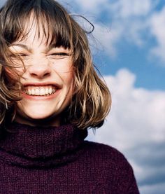 a woman with long hair smiling and wearing a purple turtle neck sweater against a cloudy blue sky