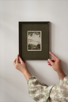 a woman holding up a framed painting on the wall above her head, with one hand reaching for it