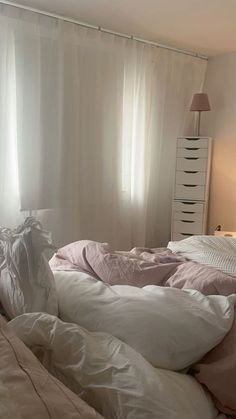 an unmade bed with white sheets and pillows in front of a window covered by sheer curtains