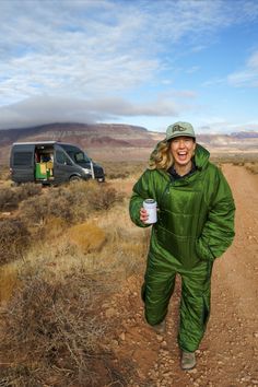 a woman walking down a dirt road holding a coffee cup in her hand and smiling at the camera