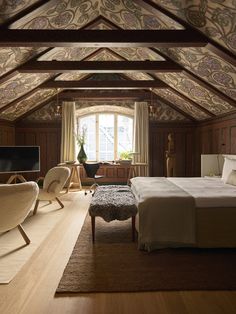 an attic bedroom with wood paneling and white bedding, two chairs, a large rug on the floor and a flat screen tv mounted to the wall