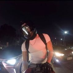 a man wearing a helmet standing next to a parked car at night with headlights on