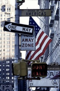 several street signs on a pole in front of tall buildings and an american flag hanging from the side