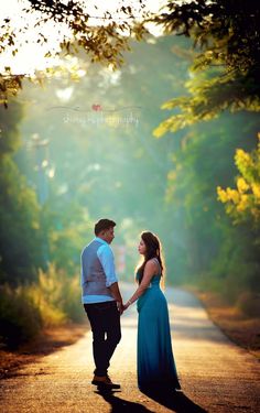 a man and woman holding hands walking down a road in the woods at sunset or sunrise