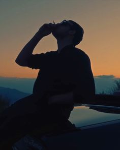 a man sitting on the hood of a car drinking from a bottle in front of a sunset