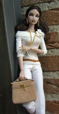 a doll holding a brown purse next to a brick wall and wearing white pants with gold accents