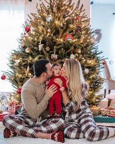 a man and woman sitting in front of a christmas tree with a baby on their lap