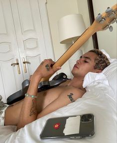 a shirtless man laying in bed with an electric guitar and cell phone next to him