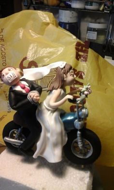 a wedding cake topper with a bride and groom on a motorbike next to a yellow plastic bag