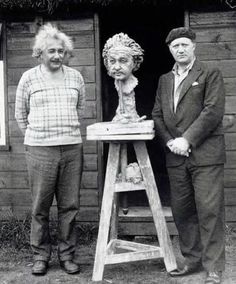 two men standing in front of a wooden sculpture on a steplade with an old man's head sticking out of it