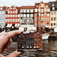 a person holding up a small model house in front of some buildings on the water