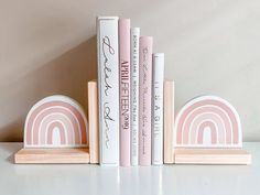 three bookends with pink and white books sitting on top of each other in front of a wall