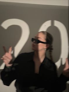 a woman in black shirt and sunglasses making the v sign with her hands while standing next to a wall