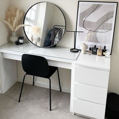 a white desk topped with a mirror next to a black chair and a vase filled with flowers